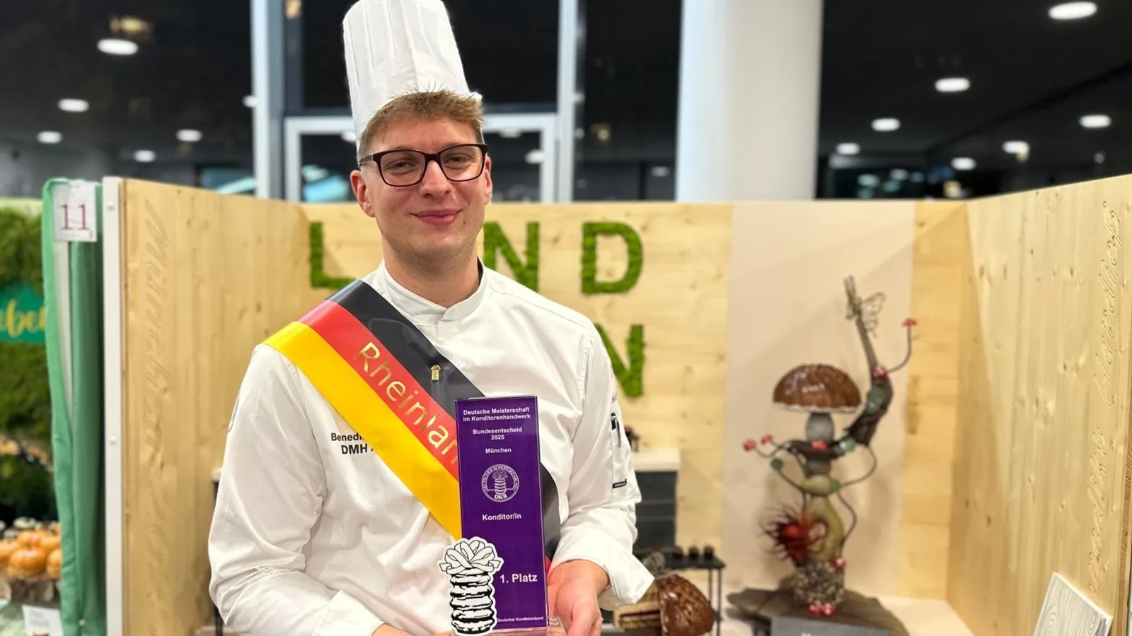 Benedikt Hardieß, Bundessieger im Konditorhandwerk, posiert vor der Kamera mit seiner Auszeichnung in der Hand.  Er trägt eine traditionelle  Berufsbekleidung.